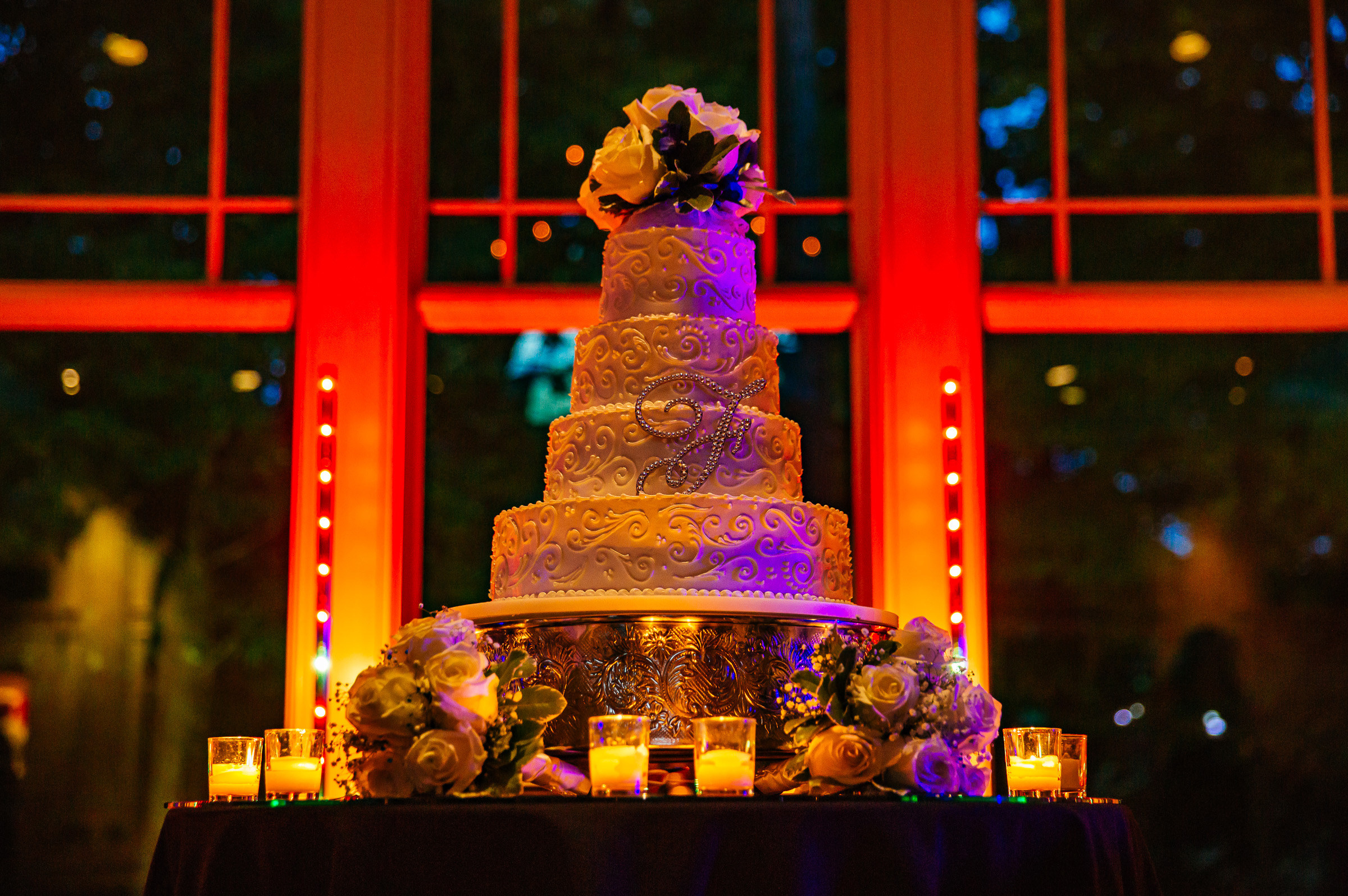 Five-tier white wedding cake featuring a rhinestone monogram letter "F" and intricate scrollwork frosting, topped with roses and displayed in front of a window illuminated by dramatic orange and red uplighting.