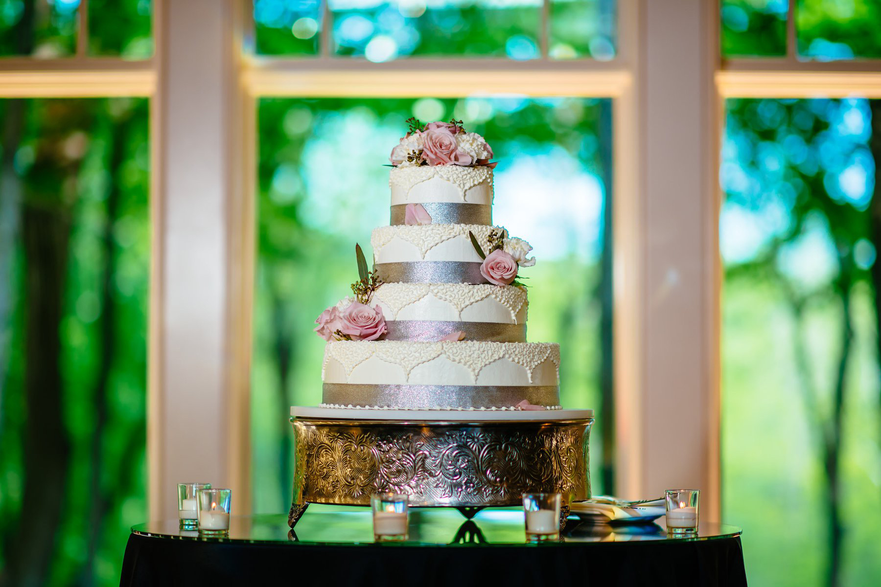 Four-tier white wedding cake with silver ribbons and pink roses, displayed in front of a window.