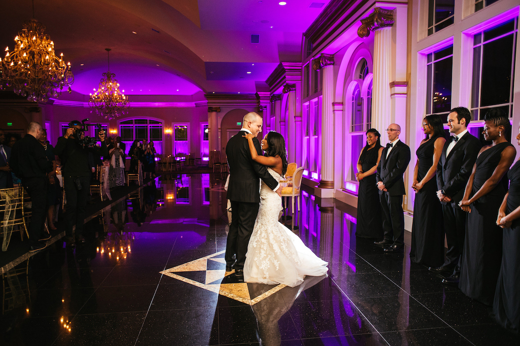 Bride and groom look at each other while sharing their first dance on a reflective ballroom floor under purple lighting, as their bridal party and guests look on.