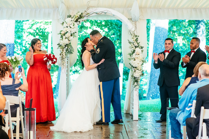Bride kisses her groom in a military dress uniform under a floral arch as wedding party applauds.