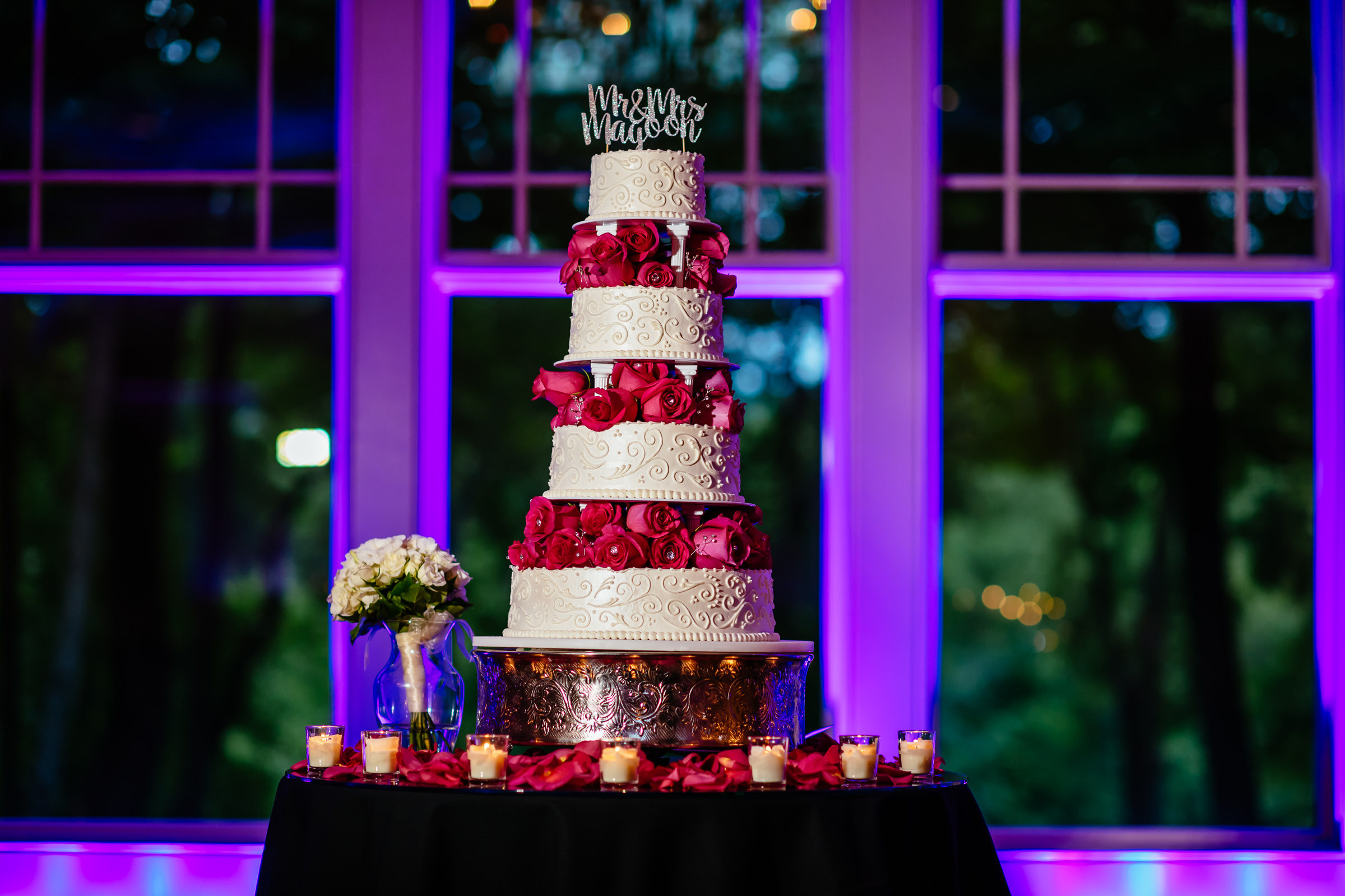 Four-tier wedding cake with red roses between layers, set against purple lighting.