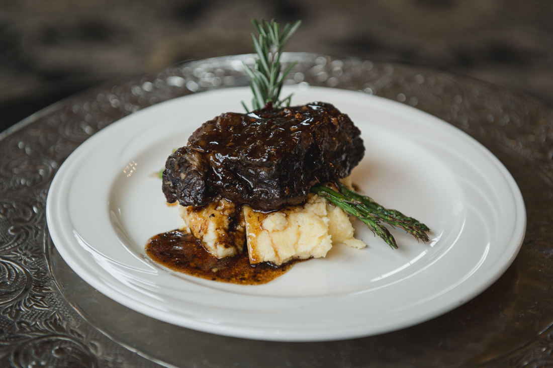 Braised short rib served over mashed potatoes with asparagus spears, drizzled with gravy and garnished with a rosemary sprig.