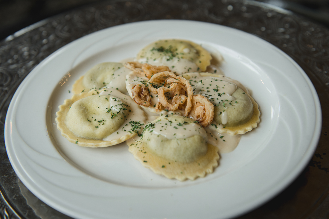 Ravioli pasta covered in a white cream sauce and garnished with fried onion straws and fresh herbs, served on a white plate.