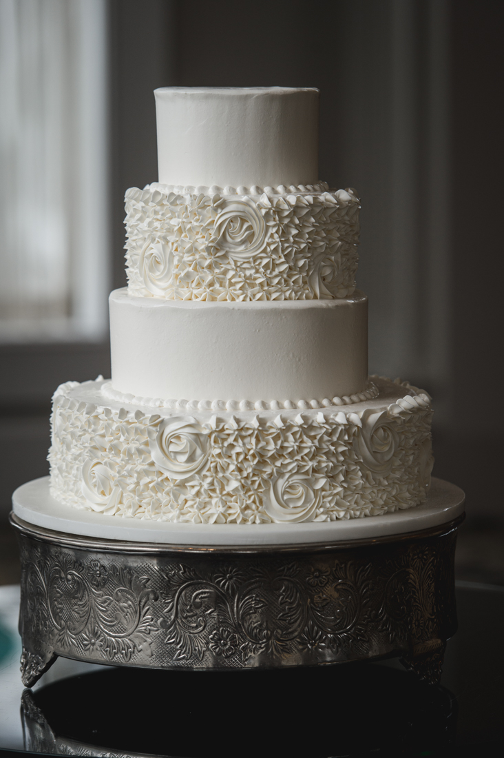 Four-tier white wedding cake with alternating smooth and rosette-textured icing on a silver stand.