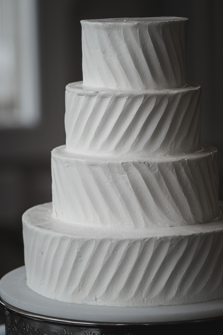 Four-tier white wedding cake with textured diagonal buttercream sweeping patterns on each layer, displayed on a silver cake stand.