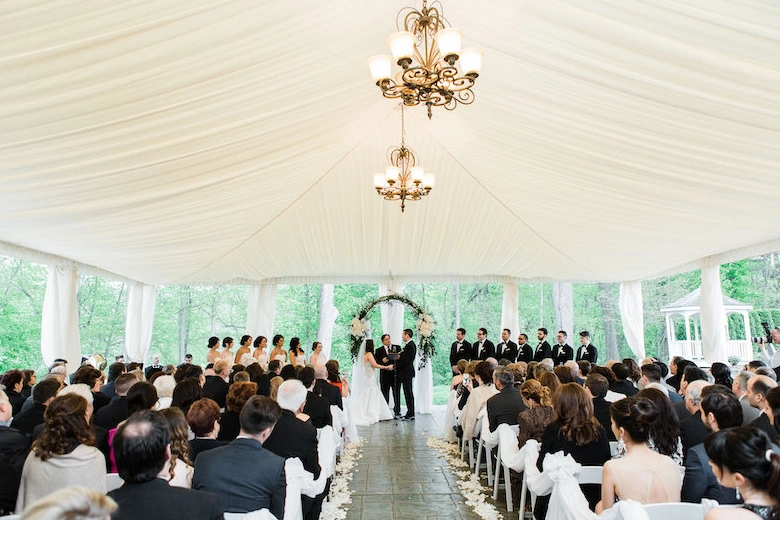 Wedding ceremony taking place under a draped white tent with chandeliers, featuring guests seated in rows facing the bride, groom, and wedding party at the altar.