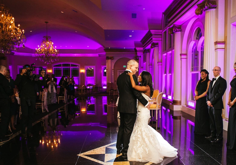 Bride and groom sharing their first dance on a glossy black dance floor in a grand ballroom illuminated by vibrant purple uplighting.