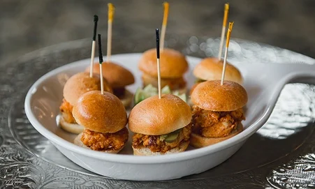 Mini fried chicken sliders with pickles, served on small brioche buns with toothpick skewers in a white dish.