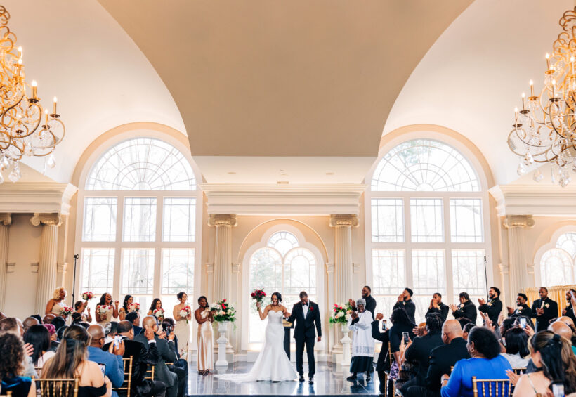 Newlyweds walk down the aisle hand-in-hand as guests cheer in a grand, sunlit ballroom.