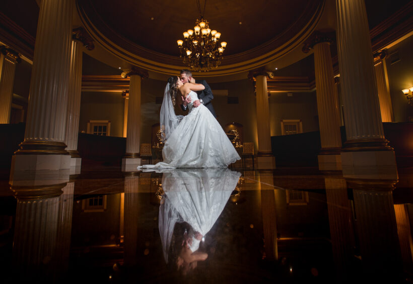 Bride and groom kiss on a shiny floor reflecting their image under a grand chandelier.