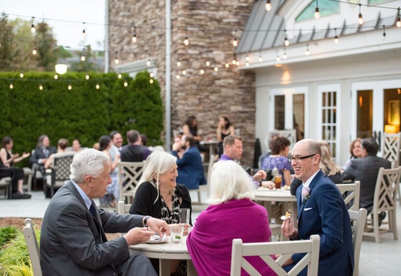 Guests laugh and talk at an outdoor wedding cocktail hour on a patio under string lights.