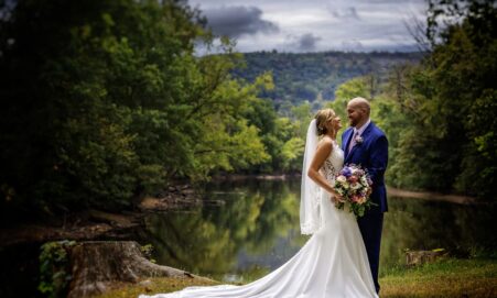 Bride and groom smile at each other by a river with a lush forest background.