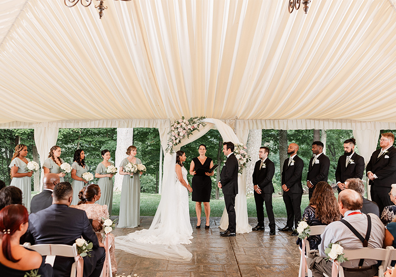 Bride and groom exchange vows under a floral arch during a tented wedding ceremony.