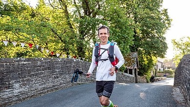 Runner smiling and wearing hydration pack crosses a stone bridge during a race, with trees and banners in the background.