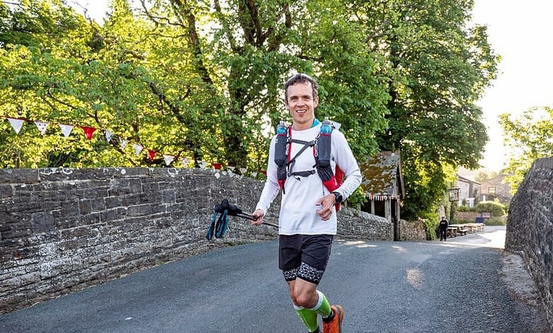 Runner smiling and wearing hydration pack crosses a stone bridge during a race, with trees and banners in the background.