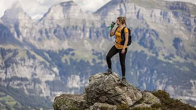 Female hiker standing on rocky outcrop drinking from a bottle, wearing yellow backpack and outdoor gear with expansive mountain range in the background.