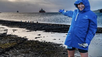 Bright female runner in waterproof jacket pointing towards the ocean on a rugged coastline during a rainy run in outdoor weather.