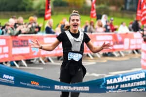 Excited female runner crossing the finish line at a marathon, celebrating success with a big smile, amidst cheering spectators and branded event banners.