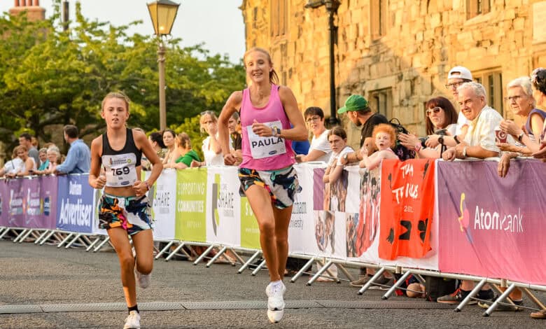 Female runner and young girl participating in a road race, showcasing running performance and athleticism; crowd supporting runners alongside historic city architecture; ideal for running insights and race analysis.