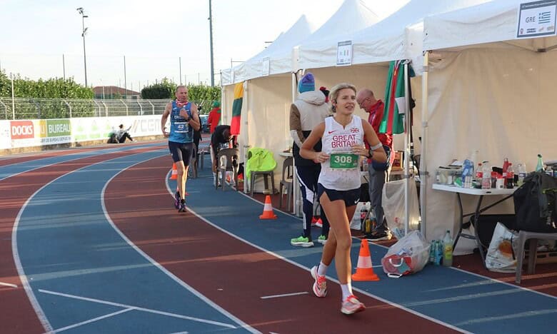 Athlete running on track at outdoor marathon event with Great Britain vest, race bib, and aid station background, highlighting endurance and competitive running.