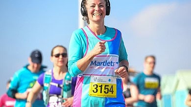 A woman running in a marathon wearing headphones, with a bright smile and a bib number 3154, showcasing endurance and running expertise, under a clear blue sky.
