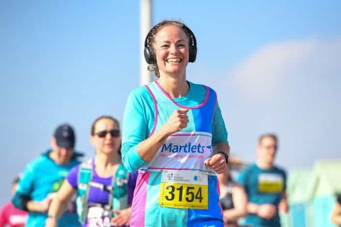 A woman running in a marathon wearing headphones, with a bright smile and a bib number 3154, showcasing endurance and running expertise, under a clear blue sky.
