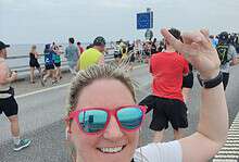 Bright smiling female runner taking a selfie during a marathon or outdoor running event on a scenic coastal route with a large crowd of participants, reflecting running insights and marathon training outdoors.