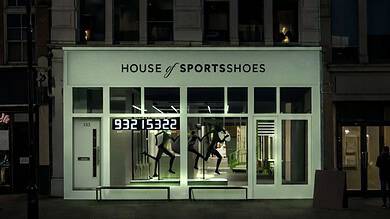 Exterior of House of Sportsshoes storefront at night with illuminated sign and mannequins inside.