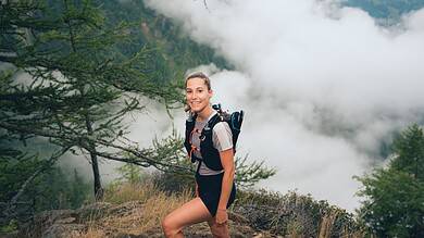 An adventurous woman trail running through lush forest terrain, showcasing ASICS trail shoes designed for outdoor running and exploration.