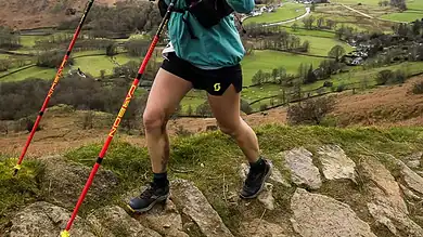 Female trail runner navigating rocky terrain in a scenic mountain landscape with lush green valleys and rolling hills in the background.
