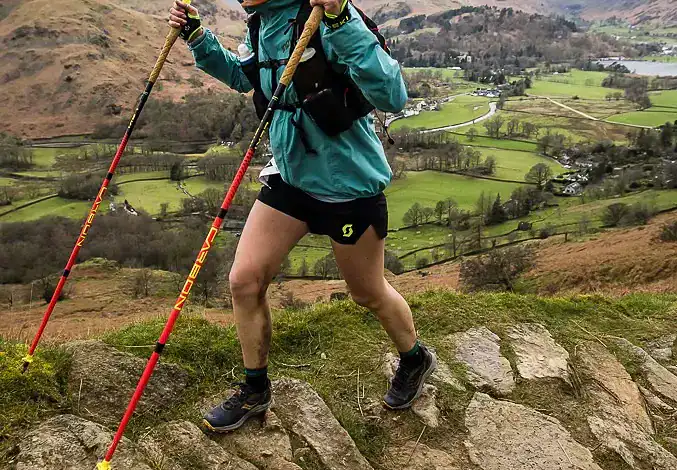 Female trail runner navigating rocky terrain in a scenic mountain landscape with lush green valleys and rolling hills in the background.