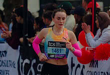 Female marathon runner in race, wearing pink arm sleeves and running shoes, with bib number 1461, competing in a city marathon event.
