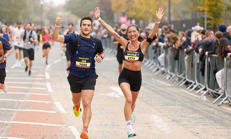 Two runners celebrating crossing the finish line at the Manchester Half Marathon, with cheering spectators and autumn trees in the background.