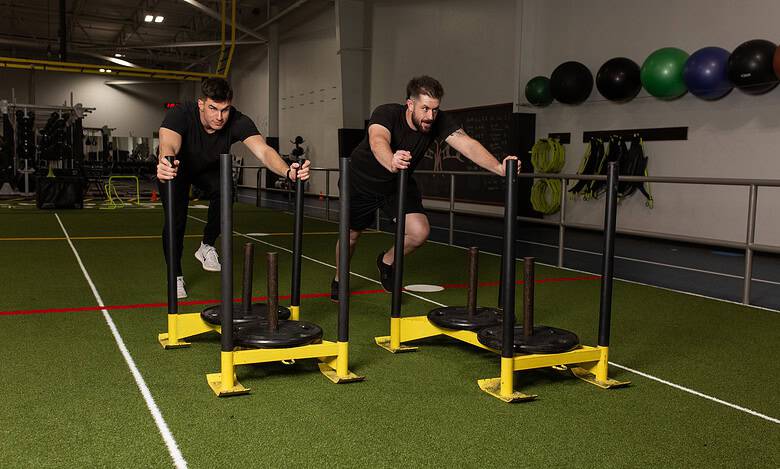Two men performing sled push exercises in a gym, focusing on strength and endurance training for runners.