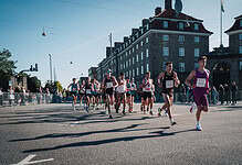 Running race with multiple athletes competing on a city street during daylight, with spectators and historic buildings in the background.