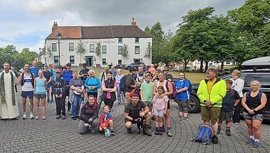 Running group gathering outdoors in a town square, diverse participants with backpacks and sportswear, some with dogs, preparing for a run or walk, under a partly cloudy sky.