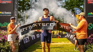 Triathlon athlete crossing the finish line at a race event, with supporters and event banners in the background, celebrating achievement and endurance.