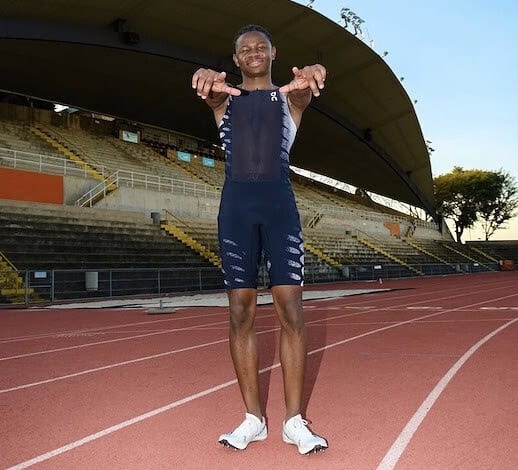 Running athlete smiling on a track at an outdoor stadium, wearing sports gear, ready for training or competition, with a clear blue sky and stadium seating in the background.