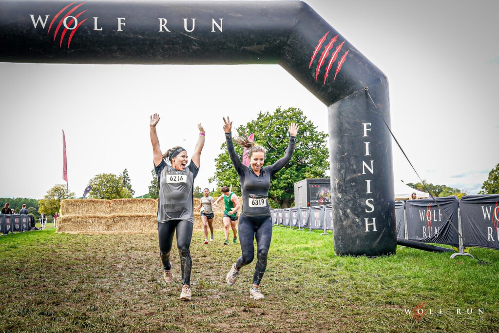 Two female runners celebrating as they cross the finish line at Wolf Run's 15th anniversary event, with a large inflatable arch and green trees in the background.