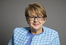 Portrait of a mature woman with short light brown hair, glasses, wearing a blue textured blazer and top, smiling against a neutral grey background.
