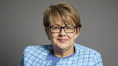Portrait of a mature woman with short light brown hair, glasses, wearing a blue textured blazer and top, smiling against a neutral grey background.