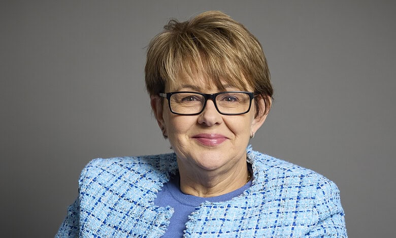 Portrait of a mature woman with short light brown hair, glasses, wearing a blue textured blazer and top, smiling against a neutral grey background.