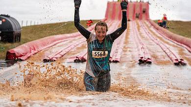 Female participant celebrating at Wolf Run obstacle course during muddy water challenge, wearing race bib 5564, with a large pink slide in the background.