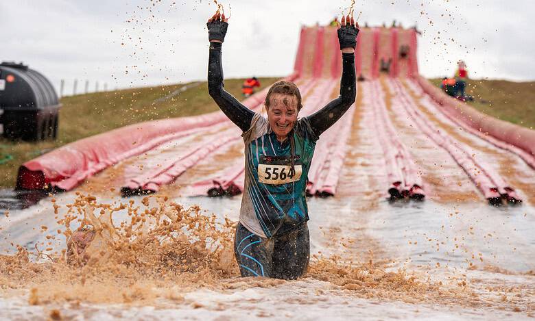 Female participant celebrating at Wolf Run obstacle course during muddy water challenge, wearing race bib 5564, with a large pink slide in the background.