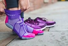 Close-up of a person putting on purple and pink running socks with purple running shoes in the background, on a city sidewalk.