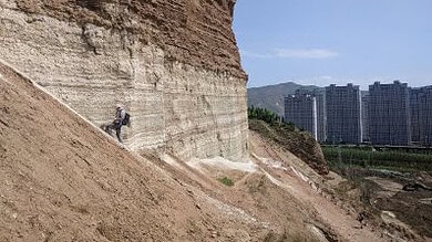 A person hiking along a steep, layered cliff face with a cityscape and mountains in the background.