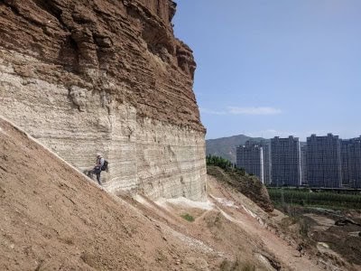 A person hiking along a steep, layered cliff face with a cityscape and mountains in the background.