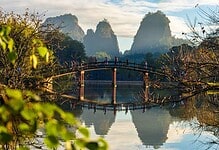 Beautiful river landscape with a wooden bridge, lush greenery, and towering karst mountains in the background, ideal for outdoor and travel enthusiasts.