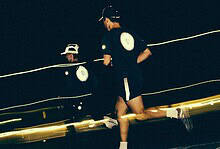 Two runners running at night on a track, illuminated by headlamps and reflective gear, with a dark background and motion blur.