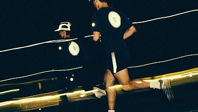 Two runners running at night on a track, illuminated by headlamps and reflective gear, with a dark background and motion blur.